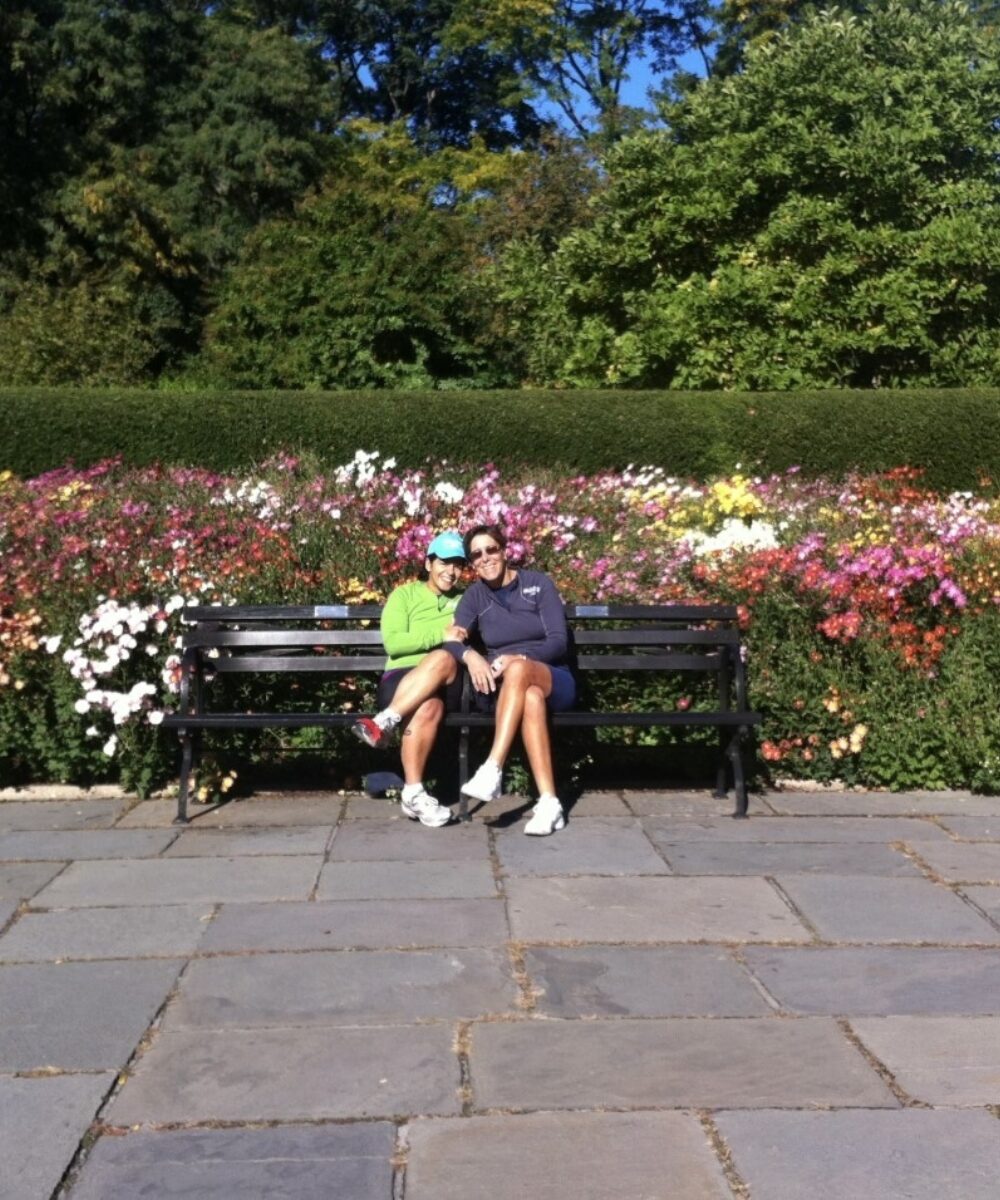 Martha and Serena sitting on a Park bench in front of colorful flowers.