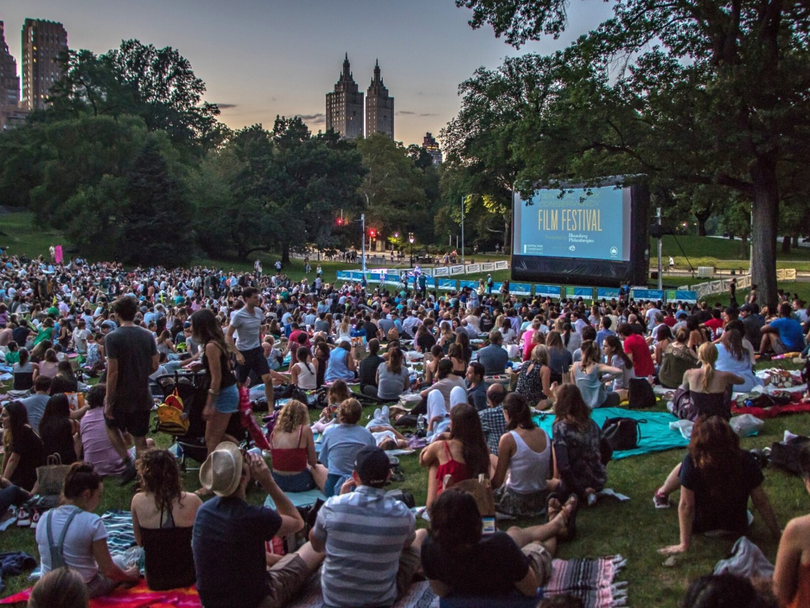 Hundreds of movie fans sit before a crowd-sized movie screen in the Sheep Medow at dusk.