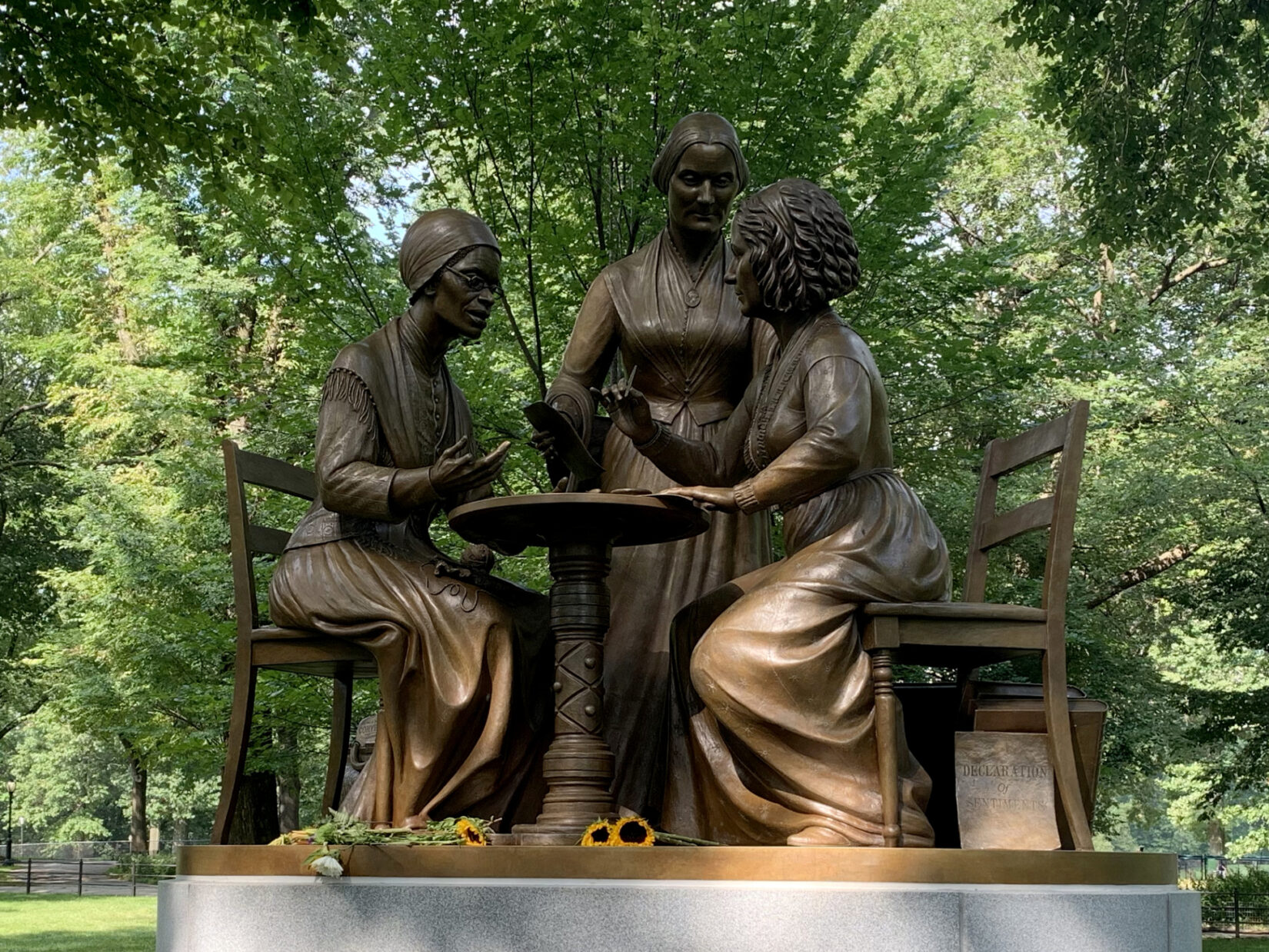 The statue is pictured under the shade of an American Elm with a few flowers left on its base