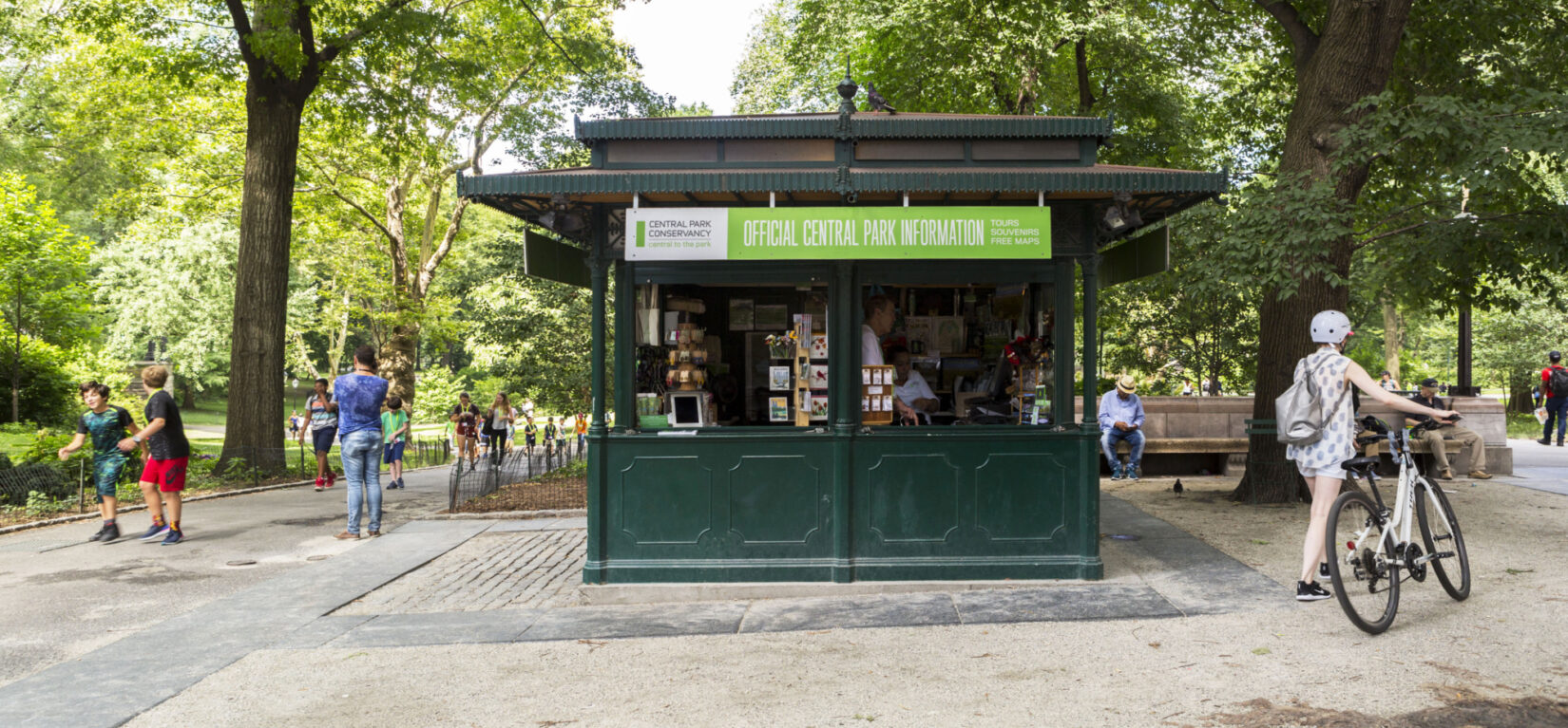The kiosk is seen framed by summer trees and passing pedestrians.