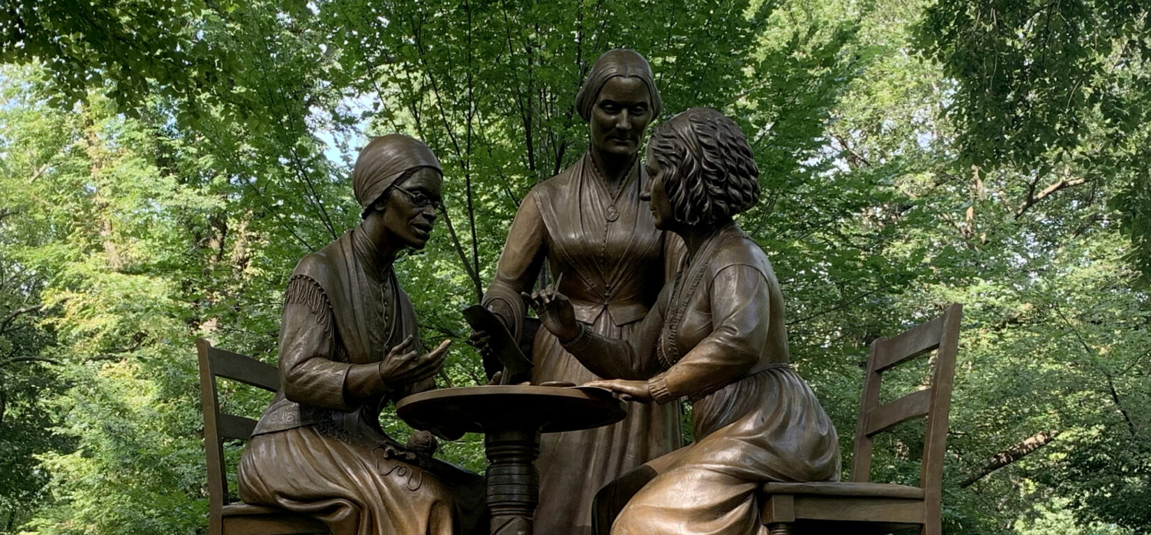 The statue is pictured under the shade of an American Elm with a few flowers left on its base