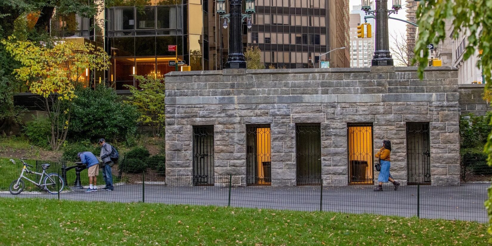 The comfort station seen in early twilight, nestled in the landscape of the Park with the buildings of on the other side of Central Park West in the background.