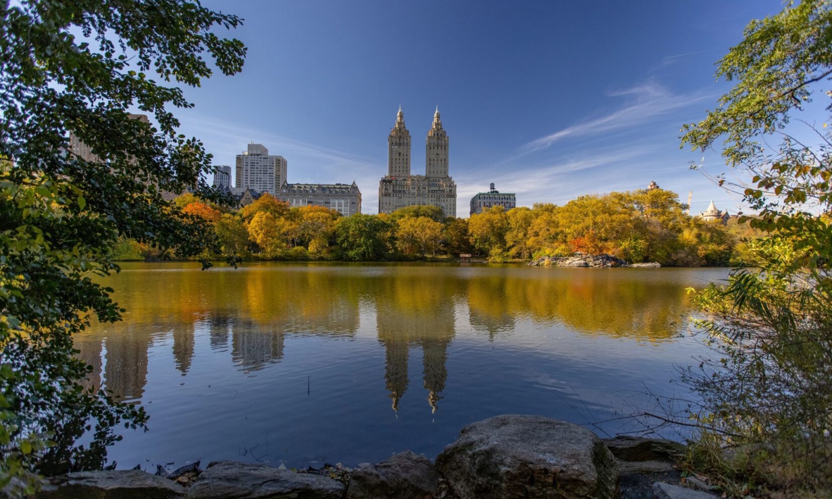 Buildings on Central Park West are reflected in still waters, viewed from the other side of the Lake in the Ramble.