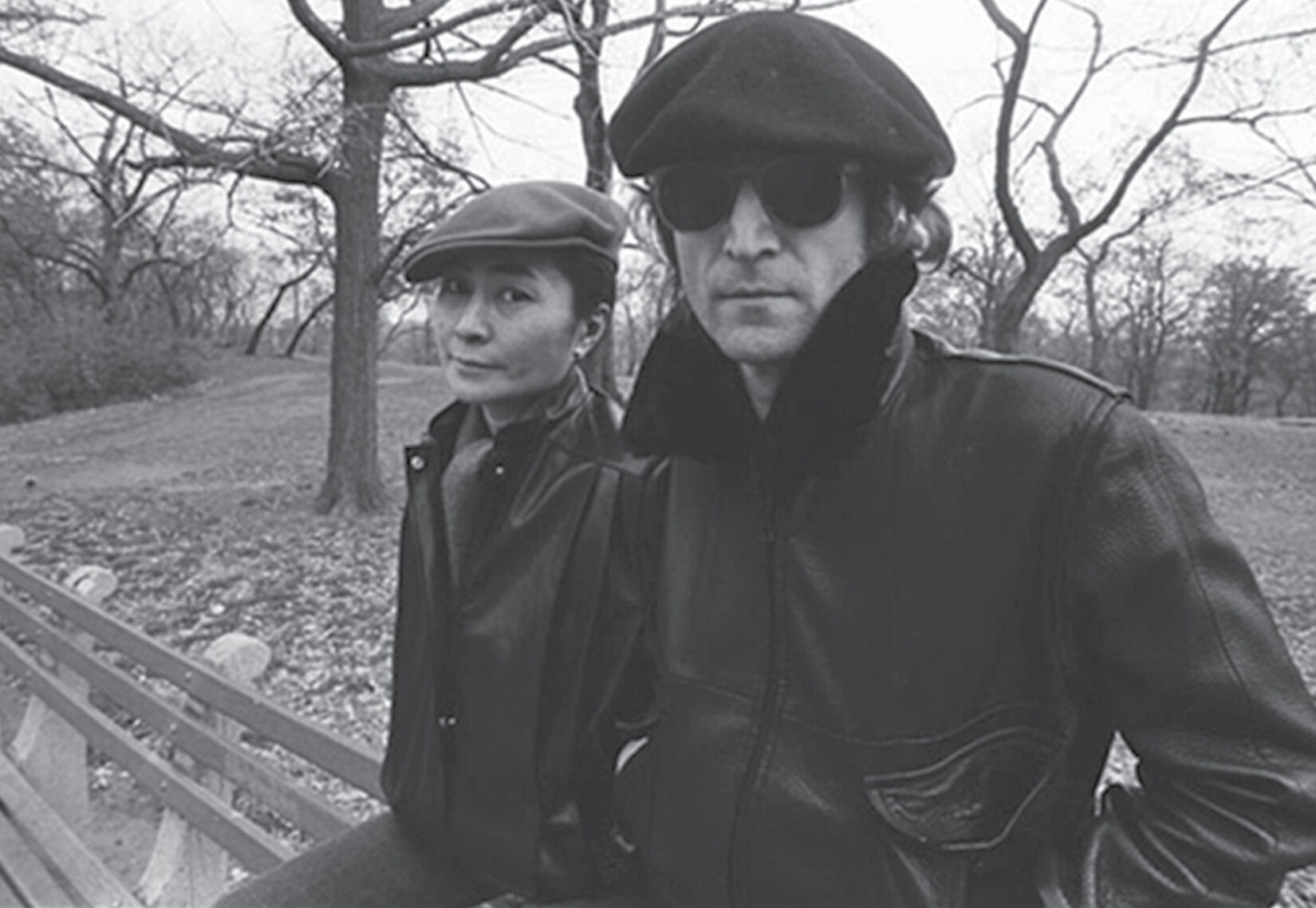 The couple are both wearing berets, sitting atop a park bench with leafless winter trees in the background.