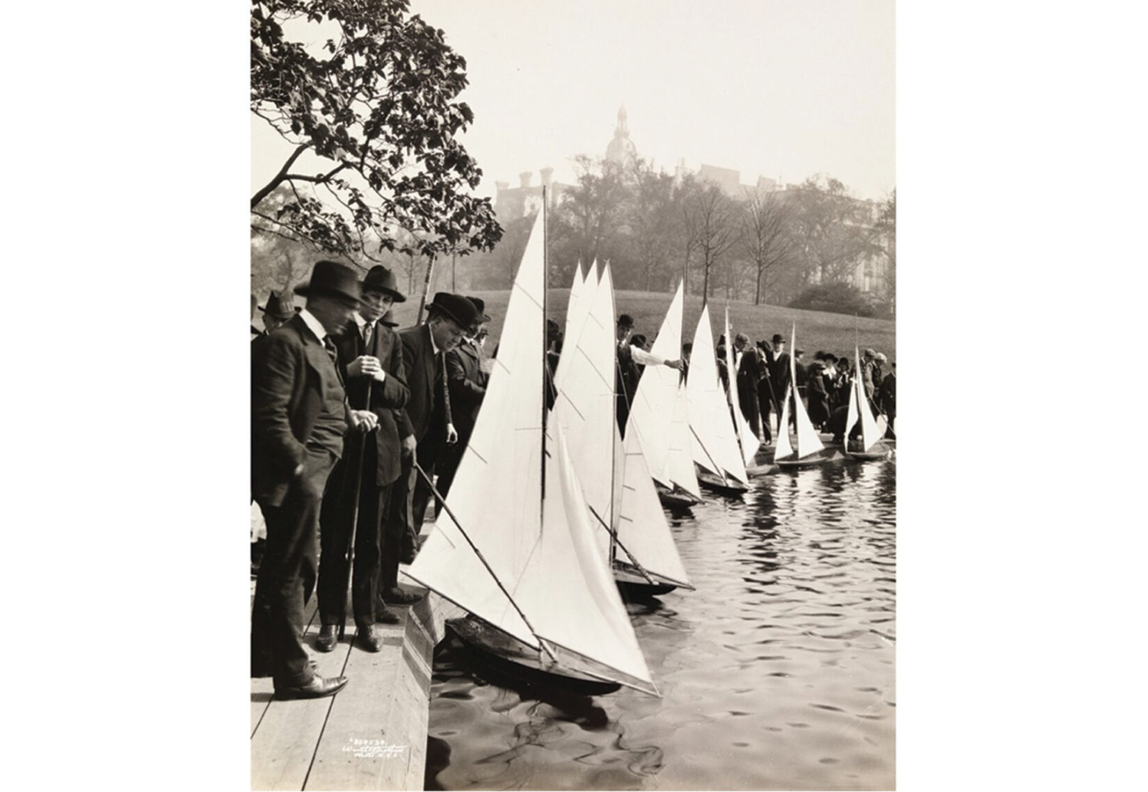 Black-and-white photo of men in suits tending model sailboats along the edge of Conservatory Water